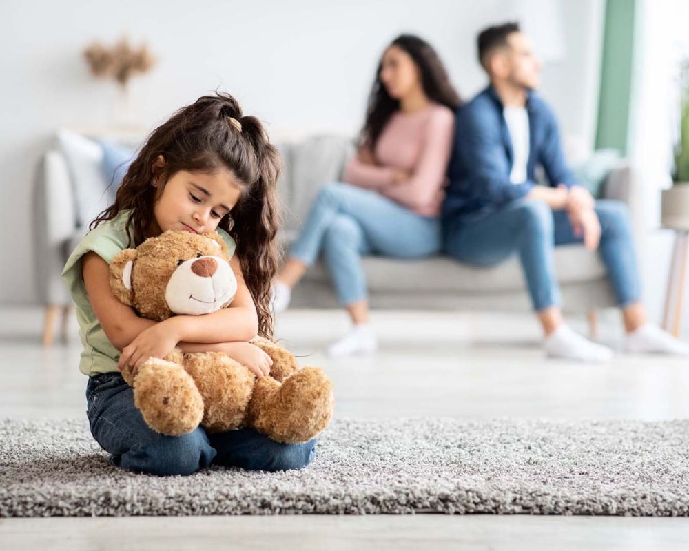 Niña abrazando un peluche con sus padres discutiendo en el fondo, simbolizando conflictos familiares, divorcios y custodia de menores.
